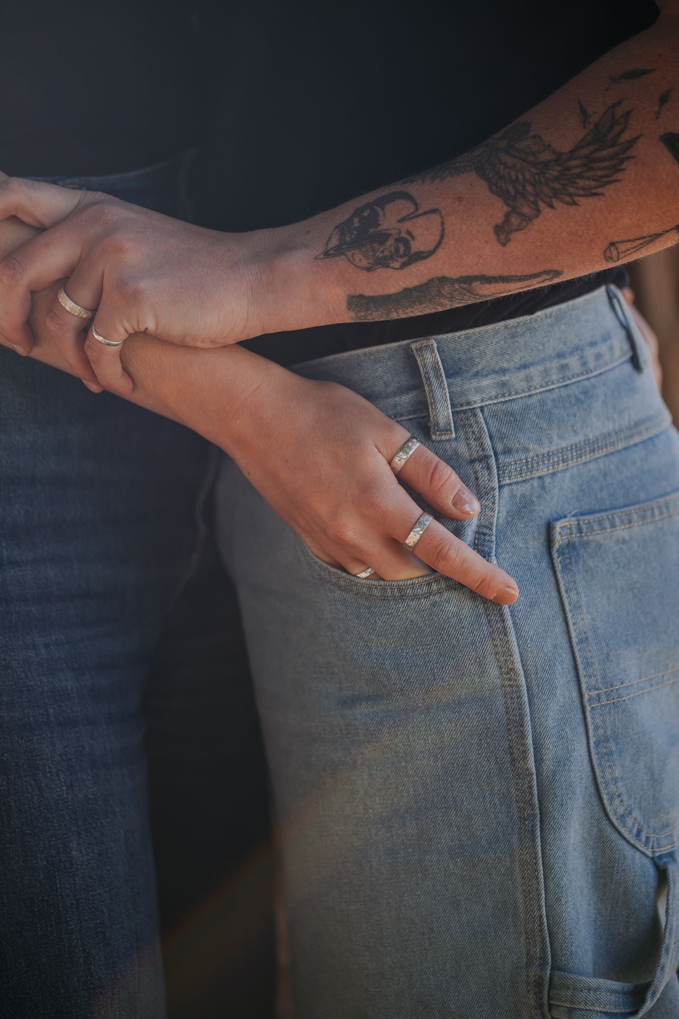 hand with silver rings put in light blue denim jeans. Another arm with tattoos holding on to the otherpersons wrist with silver jewellery.